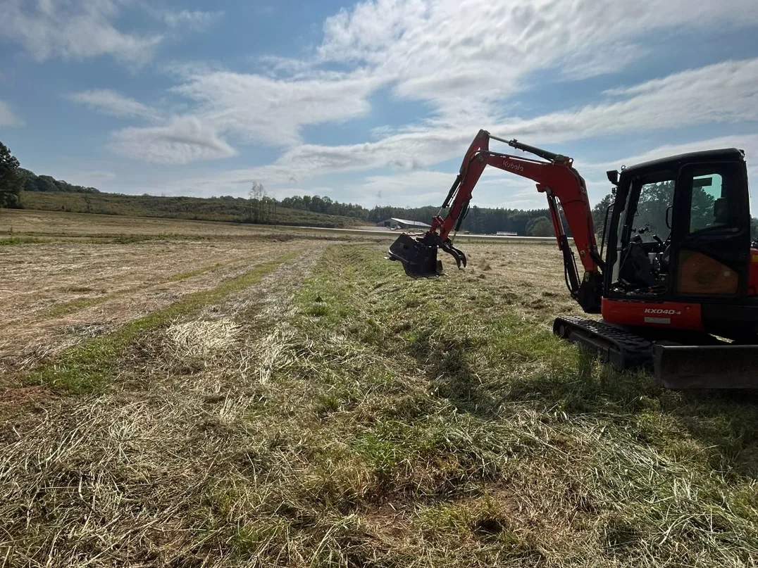 Mini excavator at a cleared ditch line across a hay field, excavation