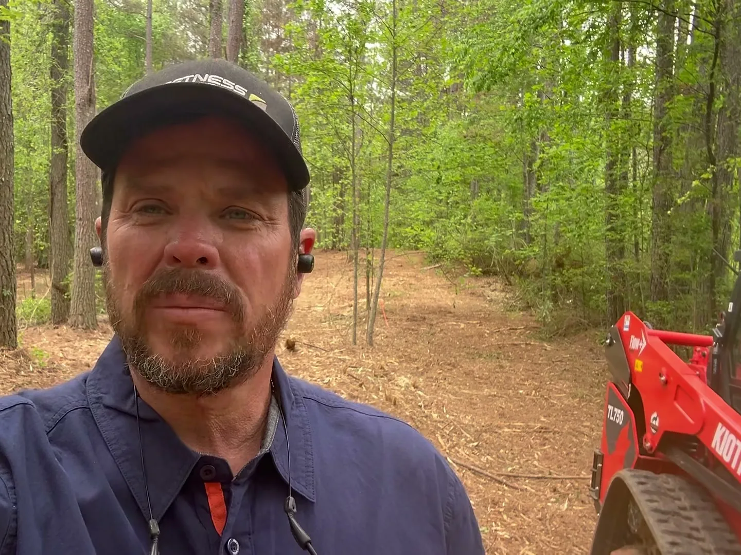 Selfie in a cap beside the red track loader on a mulched trail through the woods
